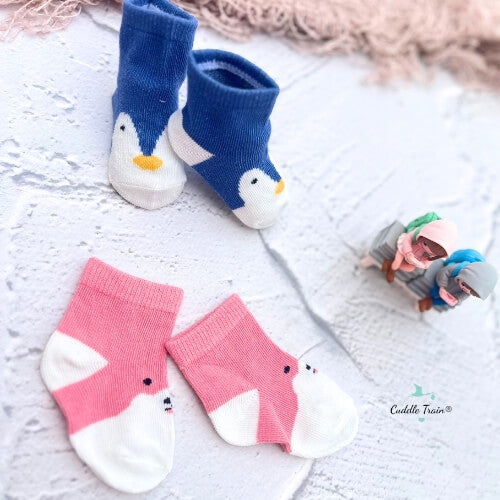 Pair of blue penguin socks and pink shark socks on a textured surface with a small toy in the background.