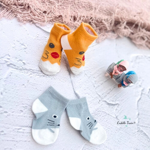 Two pairs of baby socks, one orange with animal faces and one gray with a cat face, on a textured surface.